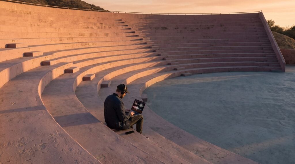 Man in headphones making a beat on a laptop, sitting alone on the steps of an empty outdoor amphitheater at golden hour