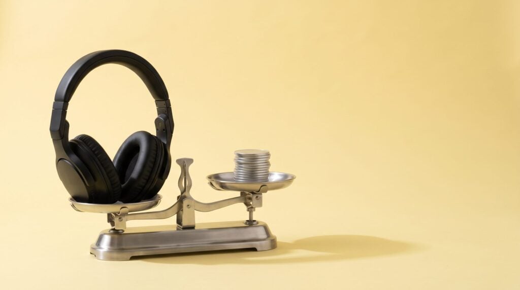 Steel balance scale weighing black studio headphones against a stack of silver coins on a butter-yellow backdrop