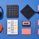 Flat lay on a cobalt blue surface: studio headphones, a drum pad controller, a graduation cap, a notebook with a hand-drawn song map, a cassette and blank coral cards