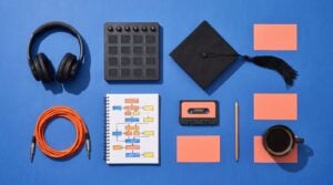 Flat lay on a cobalt blue surface: studio headphones, a drum pad controller, a graduation cap, a notebook with a hand-drawn song map, a cassette and blank coral cards