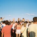 A crowd at a sunny open-air summer festival seen from behind, arms raised, singing along under a clear blue sky