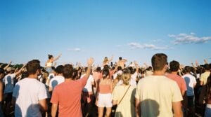 A crowd at a sunny open-air summer festival seen from behind, arms raised, singing along under a clear blue sky