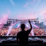 Seen from behind, a DJ raises his arm over the decks on a festival stage as the drop hits, the crowd jumping under pink and baby-blue confetti at dusk