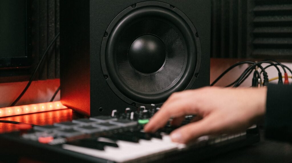 A subwoofer pumping bass in a home studio lit by a red LED strip while a producer plays a MIDI keyboard