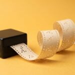 A black hand-crank music box feeding a long strip of punched paper whose holes form a repeating pattern, on a saffron-yellow backdrop