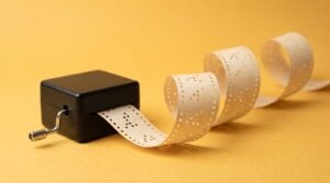 A black hand-crank music box feeding a long strip of punched paper whose holes form a repeating pattern, on a saffron-yellow backdrop