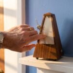 A man's hand with a wedding band starting a wooden metronome swinging, a sign it's not too late to learn music production