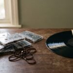 A pile of scuffed cassette tapes with tangled tape next to a glossy vinyl record sliding out of its sleeve, the gap between a rough home demo and a finished record