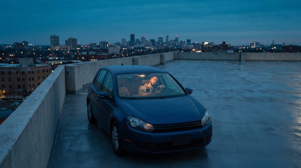 A man sits alone in his parked car on an empty rooftop at dusk, head tilted, listening closely to his beat on the car speakers
