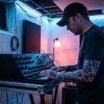 A man in a black cap with tattooed forearms plays a bass line on an analog synthesizer in a dark basement studio lit blue, with one red lamp behind him