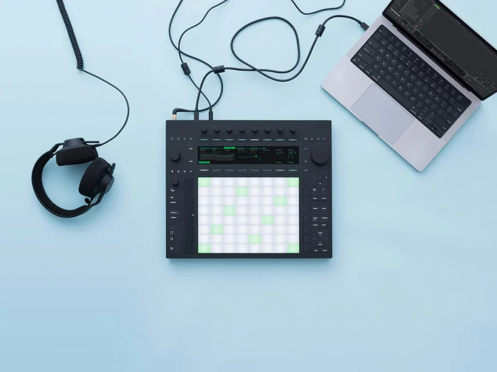 Ableton Push 3 with its 64 lit pads on a light blue desk next to a laptop and headphones, seen from above
