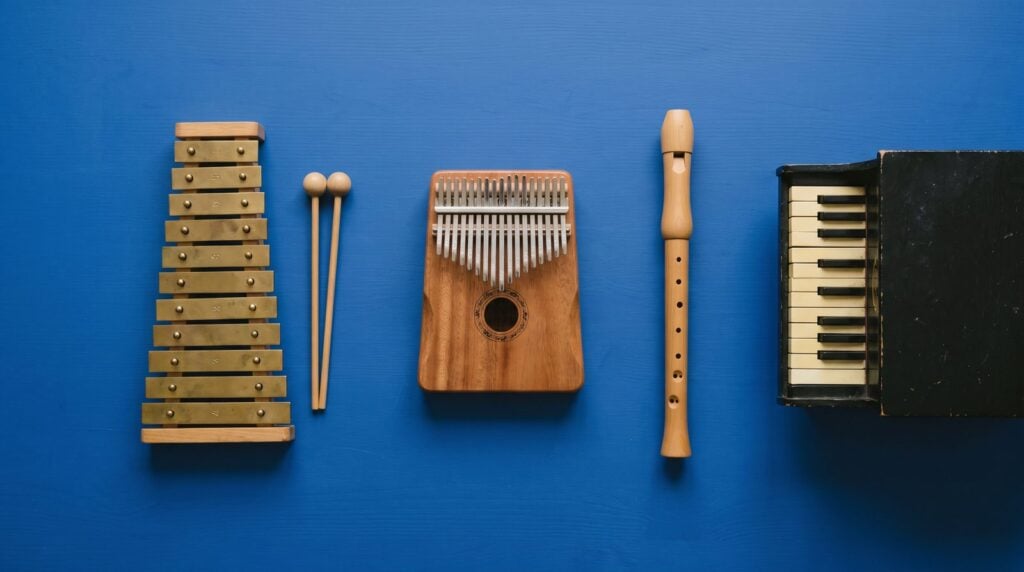 A glockenspiel with mallets, a wooden kalimba, a recorder flute and a small toy piano laid out on a blue surface, the bell, pluck, flute and key sounds trap melodies are often built from