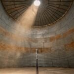 A single vintage microphone on a stand at the bottom of a huge empty steel grain silo, with daylight falling through a round opening in the roof